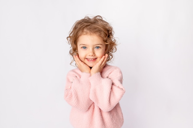 A beautiful child girl on a white isolated background in a pink sweater folded cute hands under her cheeks and smiles, space for text