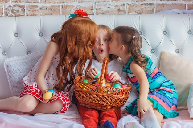 Beautiful children sit together on a bed with Easter eggs in their hands and have fun. Funny moments. Spring holidays concept. Family Easter celebration.