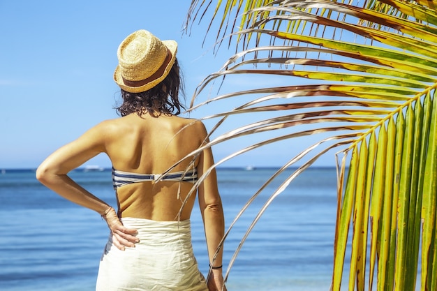 Beautiful closeup of a brunette female in a swimsuit next to a palm against blue lagoon at daytime