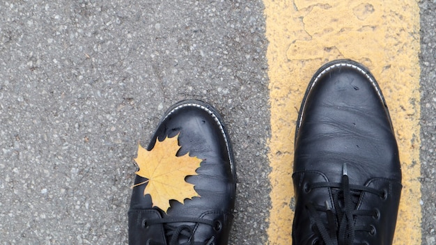 A beautiful combination of a yellow maple autumn leaf next to the yellow road markings and the legs of a girl in black boots in the park. Autumn concept, beautiful background with copy space.