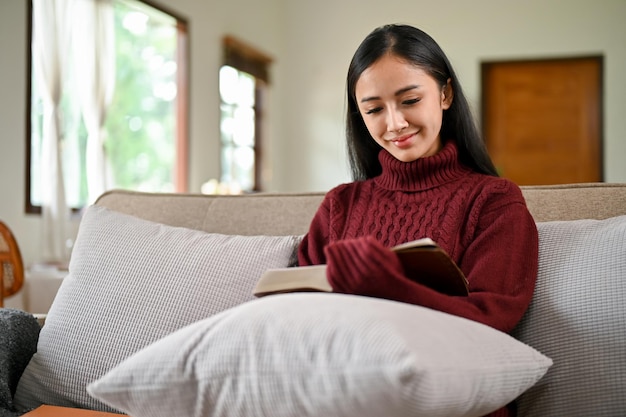 A beautiful and comforting Asian woman is reading a book on a couch in her living room