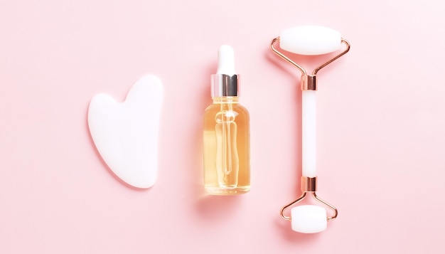 Beautiful composition of unbranded serum bottle face roller and guasha on pink background Creative trendy cosmetics flat lay with copy space