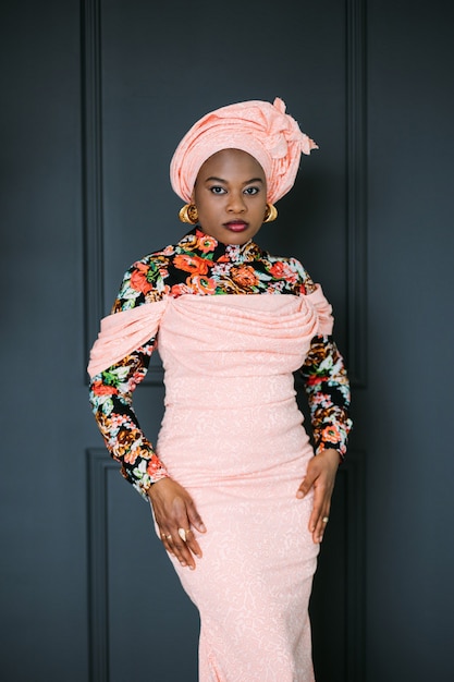 Beautiful confident African woman wearing pink stylish colorful dress and head shawl looking on camera while posing at modern dark studio background. Ethnic beauty.