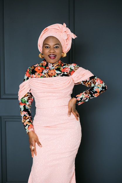 Beautiful confident African woman wearing pink stylish colorful dress and head shawl looking on camera while posing at modern dark studio background. Ethnic beauty.