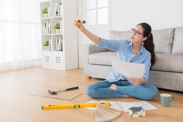 beautiful confident female house interior designer sitting in living room wooden floor and holding engineer tape ruler checking design sketch correct.