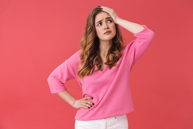 Beautiful confused young girl wearing casual clothes standing isolated over pink wall, touching head