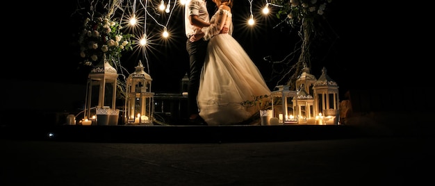 Beautiful couple is hugging near white flowers arch at the wedding ceremony Outdoors decoration Romantic rustic style Lamps lights in the evening Bride and groom together