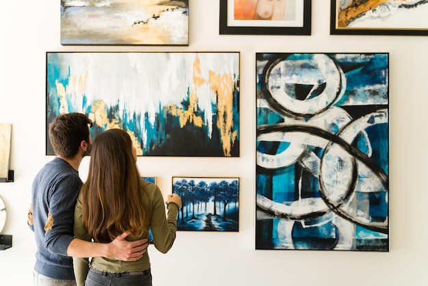 Beautiful couple seen from behind is hugging while admiring the paintings and colorful canvases hanging on the wall of the gallery