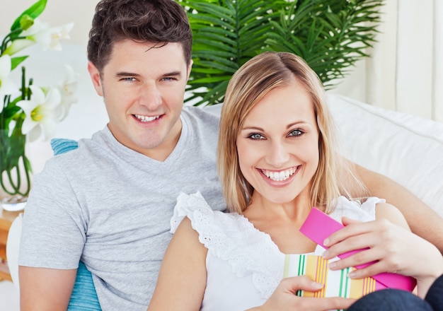 Beautiful couple smiling at the camera while woman unwrap a present lying on the sofa