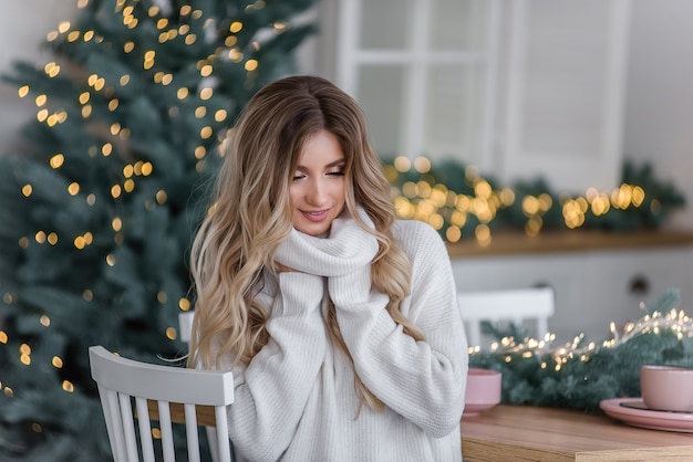 Beautiful curly blonde with long hair by the Christmas trees with garlands of lights. Close-up portrait of a girl with a white smile in a gray knitted sweater with a large collar. Copy space, texture