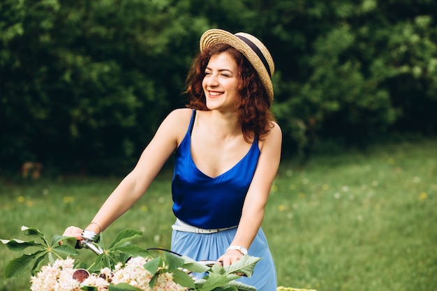 Beautiful curly-haired girl in a hat smile and holds bicycle with basket of flowers