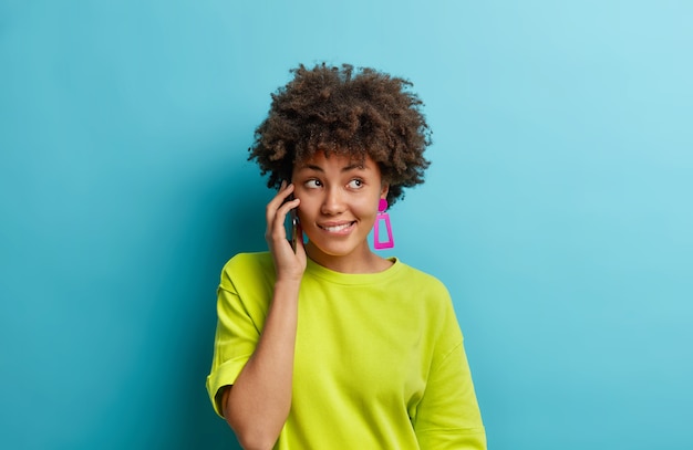 Beautiful curly haired woman talks casually with friend holds smartphone near ear bites lips gazes right enjoys good quality of device wears casual t shirt isolated on blue wall