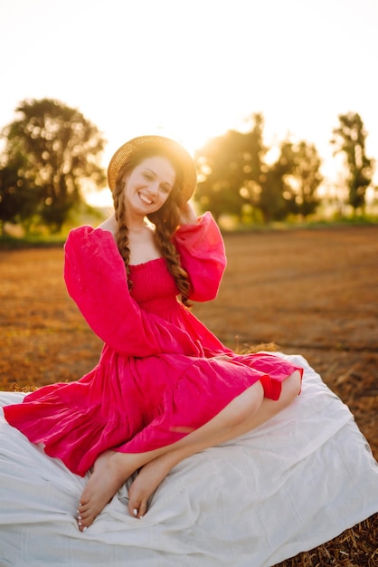 Beautiful curly woman in hat and clothes posing near hay bales in the countryside at sunset