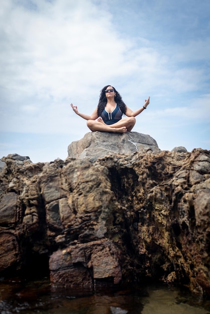 Beautiful curlyhaired woman sitting on the rock of a beach doing yoga exercise