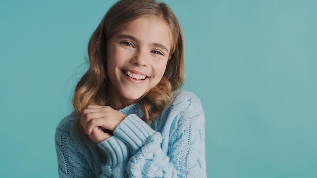 Beautiful cute blond teenage girl dressed in cozy sweater looking charming posing on camera over blue background Face expression