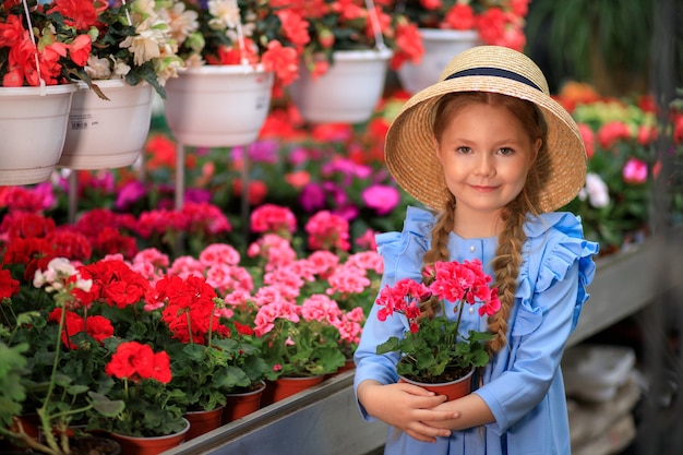 Beautiful cute girl in a straw hat holding a pink flowers pot in a greenhouse with colorful flowers