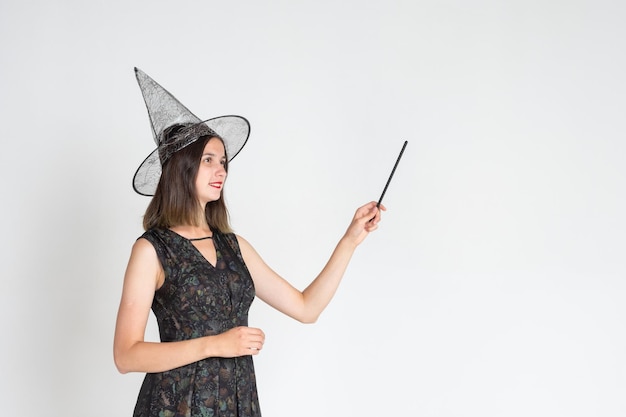 A beautiful cute girl in a witch costume wearing a hat with a magic wand shows an empty space on a white background