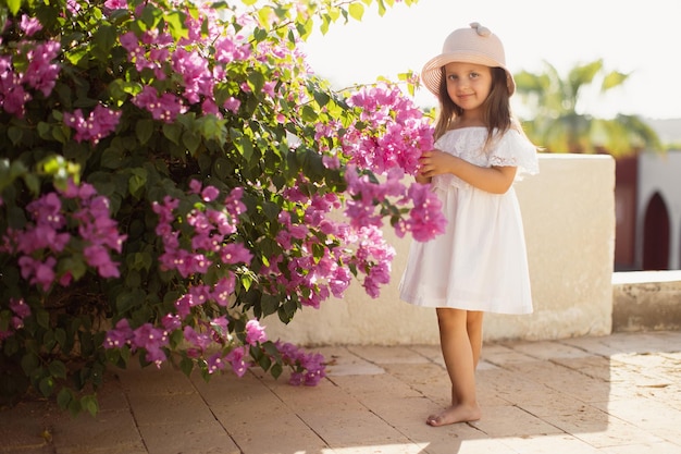 Beautiful cute girl with long blond hair in white dress and straw hat enjoying spring blooming