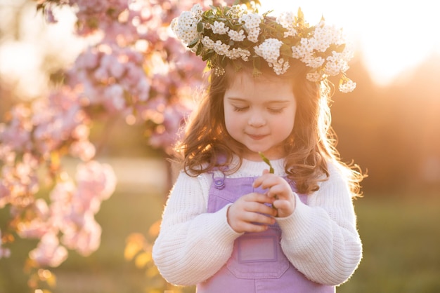 Beautiful cute kid girl wear flower wreath over sunset and blooming cherry tree