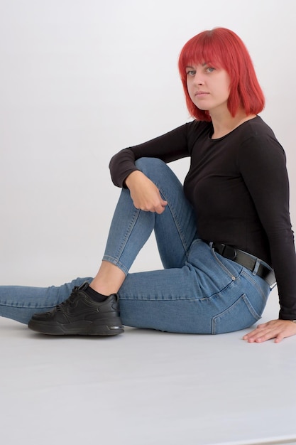 Beautiful cute young woman with orange hairstyle in jeans sitting posing in studio on white background