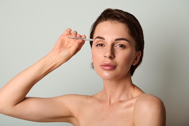 Beautiful dark haired Caucasian woman holding steel tweezers and removing hair from eyebrows posing against light gray background with copy ad space. Beauty treatment, grooming, face and skin care