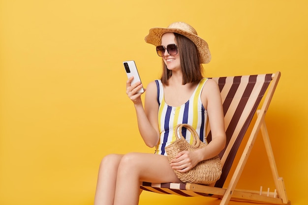 Beautiful dark haired woman addicted to modern technologies scrolls news feed via smartphone has lazy day spends summer vacations at beach wearing swimsuit