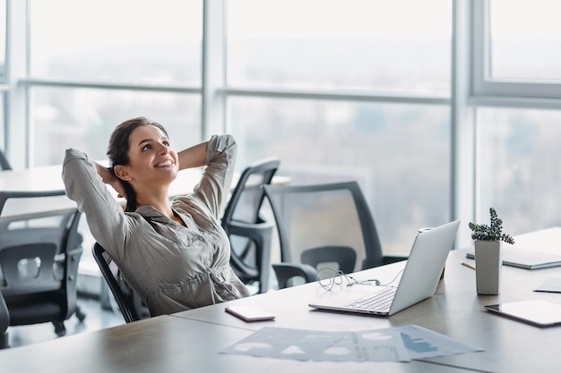 Beautiful dark-haired woman dreaming with opened eyes in office chair. Businesswoman feels relaxed after busy working day. Female leaning back in chair with hands behind head. Satisfied girl resting a