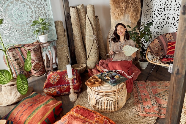 Beautiful dark haired woman in shamanic ethnic outfit in bali style decorated room is doing rappa ceremony