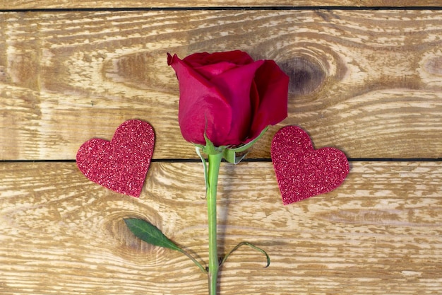 Beautiful dark red single rose and cutted glitter hearts on rough grey wooden textured background close up