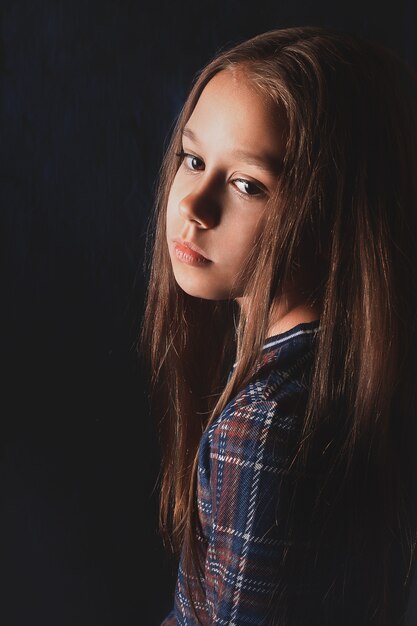 Beautiful delicate girl in plaid dress with beautiful long dark hair. Dark background.