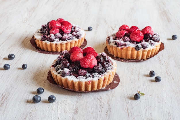 Beautiful delicious tarts, colorful pastry cakes sweets with fresh raspberries and blueberry on white wooden background.