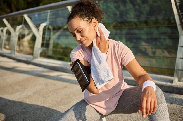 Beautiful delighted Hispanic sportswoman wiping her neck from sweat with white terry towel, sitting on squat position on the city bridge, relaxing after jog, enjoying sunny summer day during workout