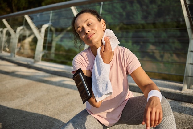 Beautiful delighted sportswoman wiping her face and neck from sweat with white terry towel, sitting on a squat position on the city bridge, relaxing after morning jog on a sunny summer day