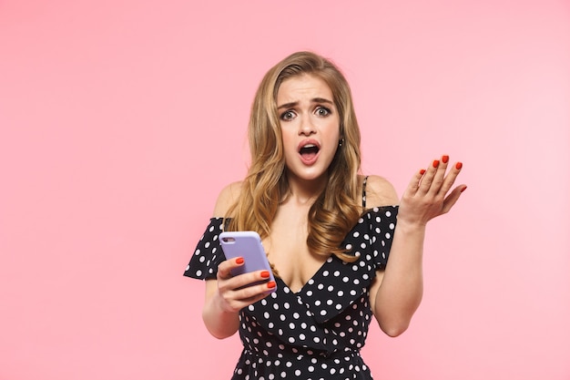 a beautiful displeased confused young pretty woman posing isolated over pink wall using mobile phone