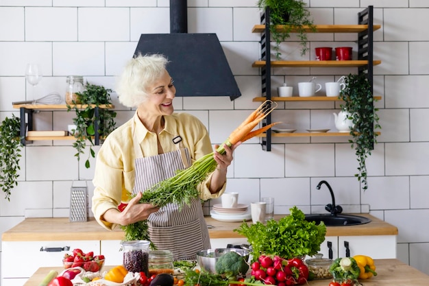 Beautiful elderly gray haired senior woman cook in cozy kitchen with fresh organic vegetables tomatoes cabbage lettuce cucumbers on table cooking healthy vegetable salad healthy food active life