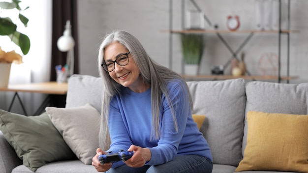 Beautiful elderly gray-haired woman playing video games using gamepads at home.