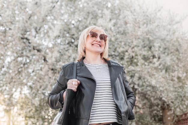 Beautiful elderly happy fashionable woman with sunglasses in fashion clothes and a leather jacket and stylish purse walks and enjoy in a park with flowering trees