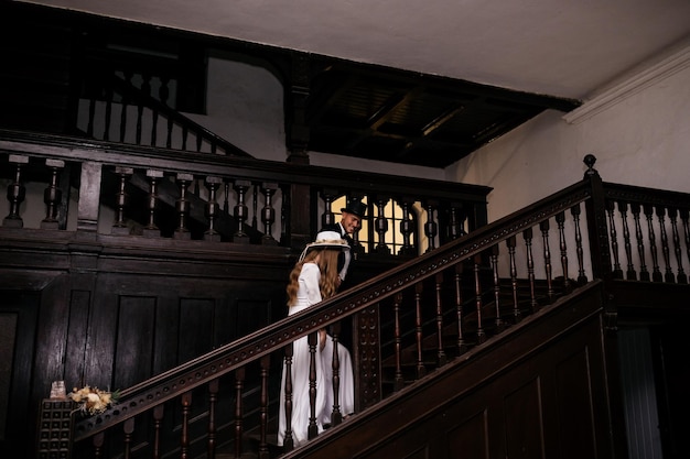 Beautiful and elegant bride in a long blue dress and hat with bouquet in hands and her handsome groom in a suit are standing in the dark room with sunlight