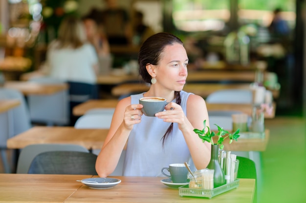 Beautiful elegant girl having breakfast at outdoor cafe. Happy young urban woman drinking coffee