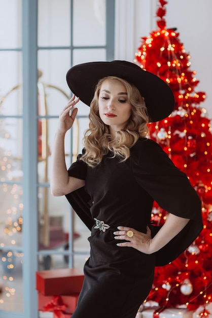 Beautiful elegant woman in a black festive dress and hat on the background of a red Christmas tree Soft selective focus