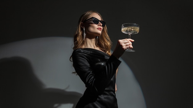 Beautiful elegant young fashion woman with sunglasses in a trendy black dress with a vintage glass of champagne on a dark background in the studio Lady celebrating a birthday at a party