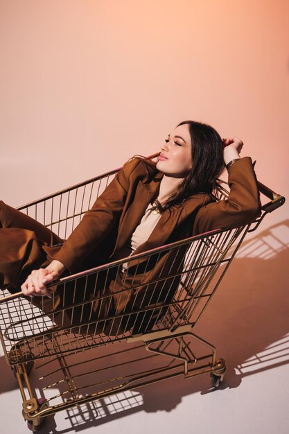 Beautiful elegant young woman model in nice stylish brown suit posing in shopping cart on gray background Young elegant woman in a suit