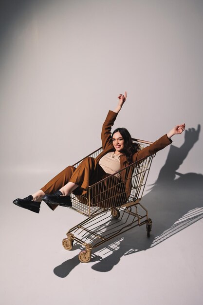 Beautiful elegant young woman model in nice stylish brown suit posing in shopping cart on gray background Young elegant woman in a suit