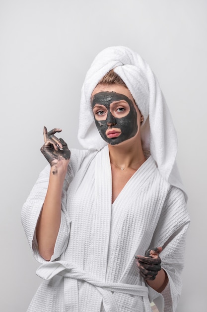 Beautiful emotional blonde posing in a white bathrobe with a towel on her head, with a clay mask and clay hands. Beauty and health concept.