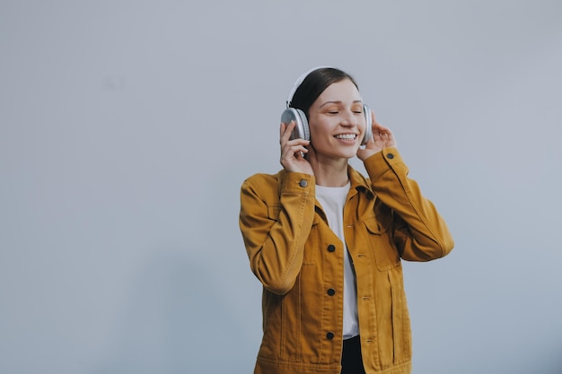 The beautiful emotional European brownhaired woman with healthy clean skin charming smile dressed in casual light clothes listens and enjoys music on headphones with tablet on a white background