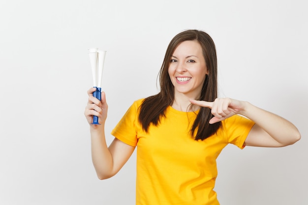 Beautiful European young happy brown-haired woman holding, blowing in football pipe, dressed in casual clothes, yellow t-shirt with shy charming smile isolated on white background. Sport fan concept.