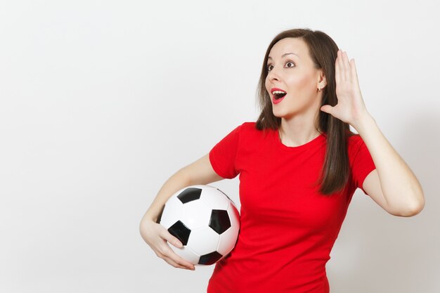 Beautiful European young woman, football fan or player in red uniform eavesdrop, hearing gesture, hold soccer ball isolated on white background. Sport play football, health, healthy lifestyle concept.