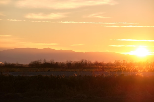 Beautiful evening panoramic landscape with bright setting sun over distant mountain peaks and asphalt road in front at sunset.