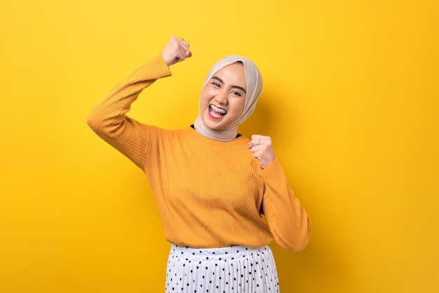 Beautiful excited Asian girl wearing hijab raising fist celebrating success isolated on yellow background