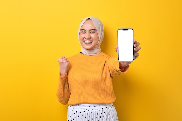 Beautiful excited Asian girl wearing hijab showing mobile phone with blank white screen raising fist celebrating good luck isolated on yellow background mockup copy space